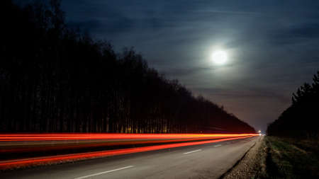 Light trails from vehicles traveling on rural road, surrounded by two rows of trees, late at night with moon on sky.の写真素材
