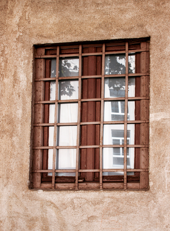 Old style window with dirty glasses and antique rusty grate with black metal bars, and red house...