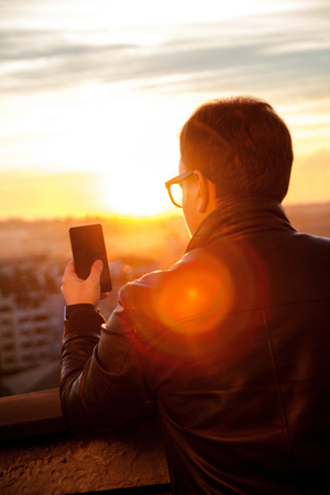 The guy standing on the roof, using smartphone and taking photos of the city skyline. Man wearing leather jacket and sunglasses using smart phone on the rooftop.の写真素材