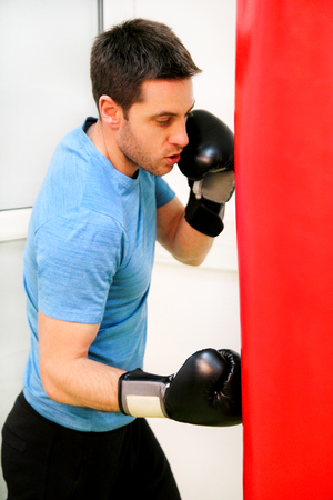Male boxer training is exercising with a punching bag at the gym club. Portrait of a young athlete handsome sports man in boxing red gloves practicing boxing on a punching bag in the sport center.の写真素材