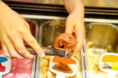 Female hand with scoop takes ice cream of chocolate from the fridge and serving in waffle cone. Woman taking scoop of tasty ice cream. Fridge with ice cream. Woman works in ice cream shop.Pastry shop.の写真素材