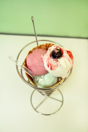 Ice Cream Holders. Strawberry, mint, vanilla with sour cherry ice cream in large waffle cone in holder, spoon. Freshly baked ice cream waffle cone sitting on silver stand. Ice cream shop. Pastry shop.の写真素材