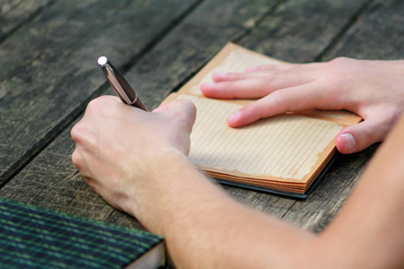 Young handsome guy sitting at wooden table, writing a book, doing homework, taking notes, learning,...