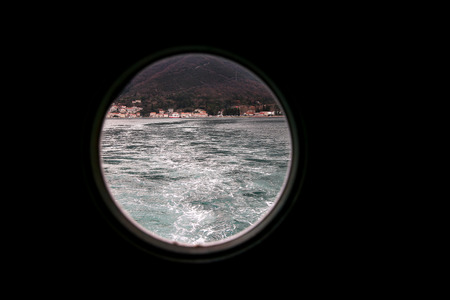 Hinged round window, storm cover on ship looking outside into Mediterranean sea. Porthole view through window on ship to turbulence made by foam of sea water from a high-speed boat on surface of sea.の写真素材