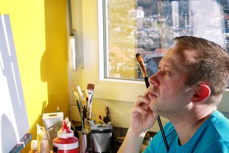 Male artist painter working in workshop with canvas on wood drawing board easel in art paint studio. Portrait of artist with paintbrush is painting on canvas with oil paints. Creativity concept.の写真素材