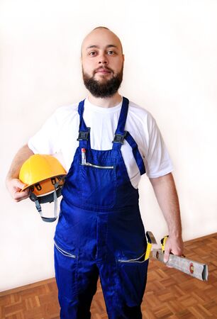 Construction worker at construction site with protective uniform, construction service tools and equipment, coverall and holds yellow helmet, hammer tool, measure tape, spirit level tool in hands.の写真素材