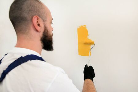 Construction worker and handyman doing finish renovation at apartment. Professional painter using paint roller brush painting of wall with yellow color on construction site. Home renovation concept.の写真素材