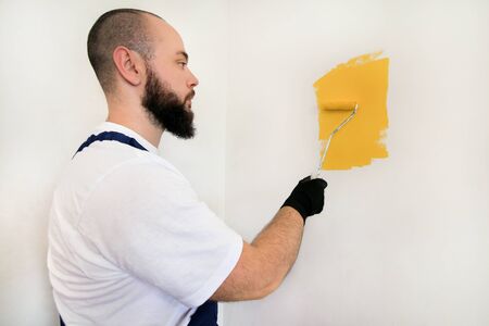 Construction worker and handyman doing finish renovation at apartment. Professional painter using paint roller brush painting of wall with yellow color on construction site. Home renovation concept.の写真素材