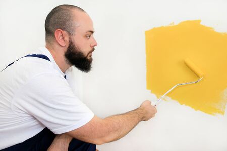 Construction worker and handyman doing finish renovation at apartment. Professional painter using paint roller brush painting of wall with yellow color on construction site. Home renovation concept.の写真素材