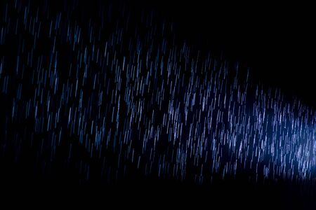 backlit raindrops cause blue streaks of light as they pass through a spotlight on a dark rainy...