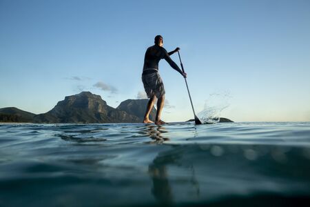 Stand Up paddle boarding on Calm ocean water, young adult surfer male standing on a surfboard and using a paddle, water surface rear view, Mount Gower and Lidgbird view, Lord Howe Islandの写真素材