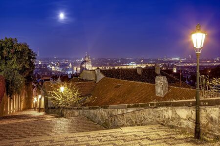 Prague city at night beautiful high view from the up sloping coblestone street, the moon in the sky and bohemian street lamps uliminate the path downhillの写真素材