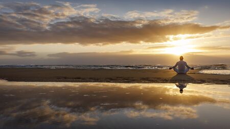 yoga meditation silhouette lotus sunrise beach, mindfulness, wellness and wellbeing concept, water reflection of man in yoga lotus pose sitting alone on sand with ocean cloud background, copy spaceの写真素材