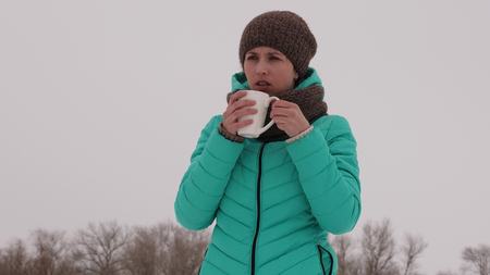 Frozen girl warms herself with hot mug in the frosty fresh air in winter. Drink tea in winter park....