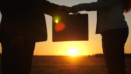 business woman and businessman make deal, woman passes briefcase to man and shakes hands, against backdrop of a beautiful sunset.の写真素材