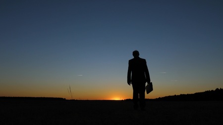 silhouette of businessman with briefcase walking across field at sunset, against a blue skyの写真素材