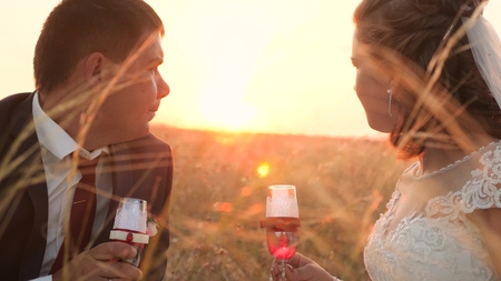 Happy bride and groom drink champagne from beautiful glasses in bright rays of sun. enamored newlyweds drink wine in meadow in rays of a beautiful sunset. happy wedding concept. close-up.の写真素材