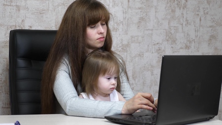 Working mother with her little daughter at the table. Busy woman working on laptop with baby on hands. Working mom with beautiful infant on hands in cozy home. Female freelance work. Modern motherhoodの写真素材