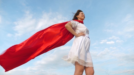sexy superhero girl standing on the field in a red cloak, cloak fluttering in the wind. girl dreams of becoming a superhero. Slow motion. young girl in a red cape dream expressionの写真素材