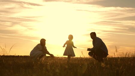 Mom and Dad playing with their daughter in park at sunset. kid takes the first steps. Happy family playing with the child in the rays of sun. baby goes from dad to mom and laughs. Slow motion.の写真素材