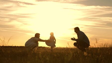 Mom and Dad playing with their daughter in park at sunset. kid takes the first steps. Happy family playing with the child in the rays of sun. baby goes from dad to mom and laughs. Slow motion.の写真素材