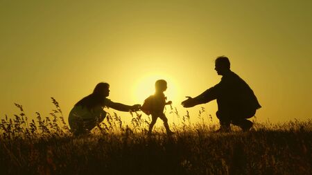 Little daughter with parents jumping at sunset. Silhouettes of mom dad and baby in the rays of dawn. Family concept. Walking with a small child in natureの写真素材