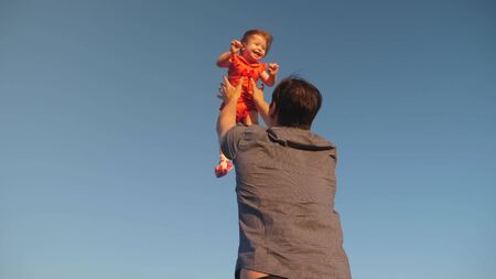Dad throws up his daughter in the blue sky. Father plays with a small child. happy family playing in the evening against the sky. Dad throws up the baby, the kid smiles. Slow motion. happy familyの写真素材
