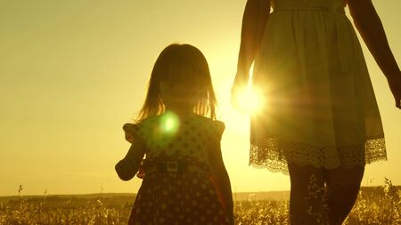 Mom walks across the field with a small child at sunset. daughter holds moms hand, a happy family walks in evening out of town. mother and baby are resting in park. child plays with mother in meadow.の写真素材