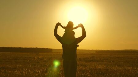 Dad carries beloved baby on his shoulders walking around field at sunset. A child with parent walks in evening at sunset of sun. little daughter riding with dad on his shoulders in park. Slow motion.の写真素材