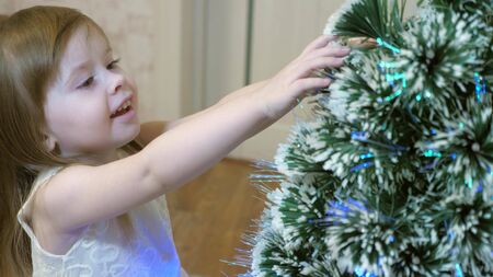little girl examines a garland on Christmas tree. child plays by Christmas tree in childrens room. beautiful artificial christmas tree . happy childhood concept. Family plays for Christmas holidaysの写真素材