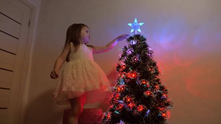 happy christmas holiday concept for children. child examines a christmas star on a holiday tree. little girl plays near a Christmas tree in a childrens room. beautiful artificial christmas tree.の写真素材