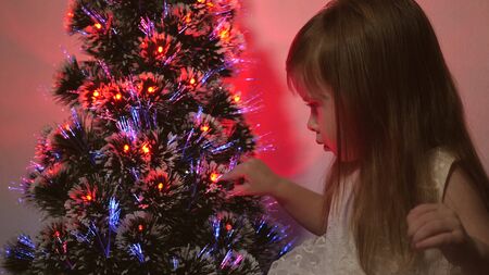 daughter examines garland on Christmas tree. small child plays by Christmas tree in children's room. beautiful artificial christmas tree . happy childhood concept. Family plays for Christmas holidaysの写真素材