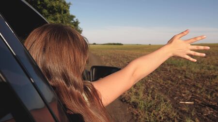happy female traveler rides car along a country road. hand from the car window catches wind. Girl with long hair is sitting in front seat of car, stretching her arm out window and catching glare sunの写真素材