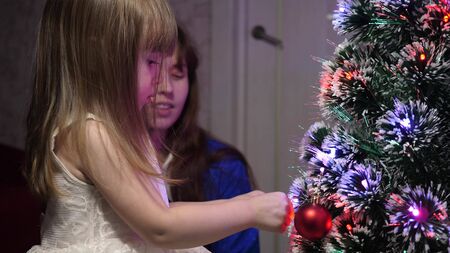 baby and mom hang a red ball toy on Christmas tree. happy childhood concept. child and mother decorate tree with christmas balls. small child and a parent are playing by Christmas tree.の写真素材