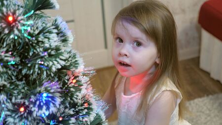 Happy little baby is playing near the Christmas tree and smiling. The girl strokes her hairy beautiful spruce. Smart spruce. New Year. Christmas Holidaysの写真素材