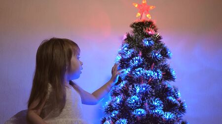 small child plays by Christmas tree in childrens room. daughter examines garland on Christmas tree. beautiful artificial christmas tree . happy childhood concept. Family plays for Christmas holidaysの写真素材