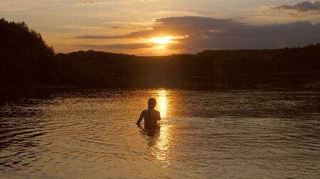 girl goes into the water at sunset the fiery sun. young woman bathes in the evening in the river. rest by the water.の写真素材