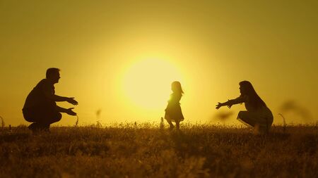 happy baby goes from dad to mom. parents are taught to walk small child, little girl makes her first steps in sun, slow motion. parents play with their little daughter.の写真素材