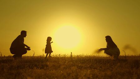 happy baby goes from dad to mom. parents are taught to walk small child, little girl makes her first steps in sun, slow motion. parents play with their little daughter.の写真素材