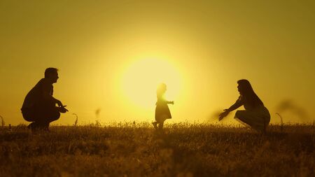 happy baby goes from dad to mom. parents are taught to walk a small child, little girl makes her first steps in sun, slow motion. parents play with their little daughter.の写真素材