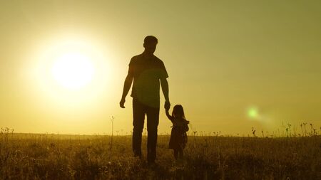 little daughter and dad walk around field holding hands. child holds father's hand. dad and baby are resting in park. child plays with his father. happy family and childhood conceptの写真素材