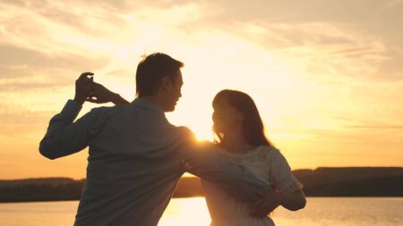 Loving man and woman dance in bright rays of sun on the background of the lake. Young couple dancing at sunset on beach. Happy guy and girl waltz in the evening in the summer park.の写真素材