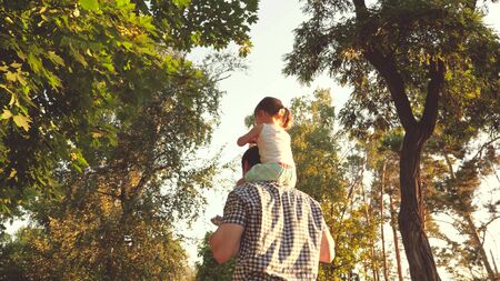 Dad carries on shoulders of his beloved child, in park. Father walks with his daughter on his shoulders under trees. child with parents walks on the day off. Happy family is relaxing in park.の写真素材