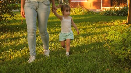 little daughter and mother holding hands are walking in park on lawn. child walks on green grass. Mother and baby rest on lawn. concept of a happy childhood. A happy family is walking with a child.の写真素材