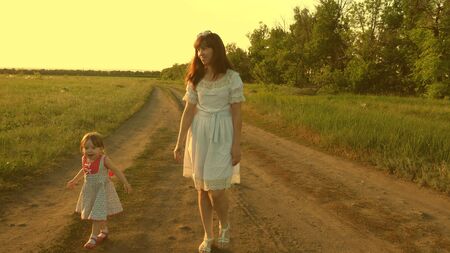 Mother and baby rest in park. concept of a happy childhood. little daughter and mom walk along road past field. child plays running away from mom. happy family with a child walks in countryside.の写真素材