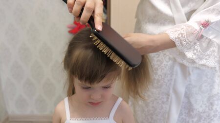 mother combs the childs hair with a comb. concept of beautiful hairstyle for daughter. a happy family. Mom Braids the Braid Of Her Little Daughter.の写真素材