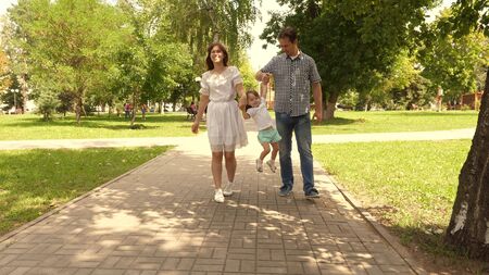 happy child plays with dad and mom holds their hands and jumps. little daughter jumping holding hands of dad and mom in the park. Family concept. Walking with small kid in nature.の写真素材