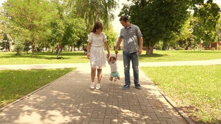 happy child plays with dad and mom holds their hands and jumps. little daughter jumping holding hands of dad and mom in the park. Family concept. Walking with small kid in nature.の写真素材