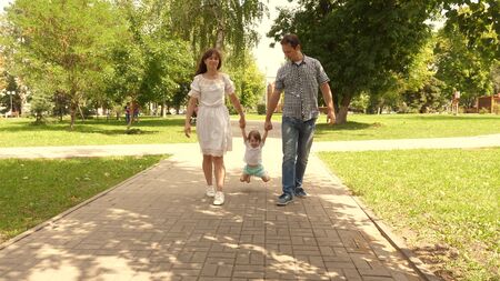 happy child plays with dad and mom holds their hands and jumps. little daughter jumping holding hands of dad and mom in the park. Family concept. Walking with small kid in nature.の写真素材