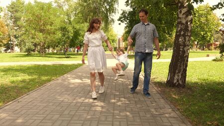 happy child plays with dad and mom holds their hands and jumps. little daughter jumping holding hands of dad and mom in park. Family concept. Walking with small kid in nature.の写真素材